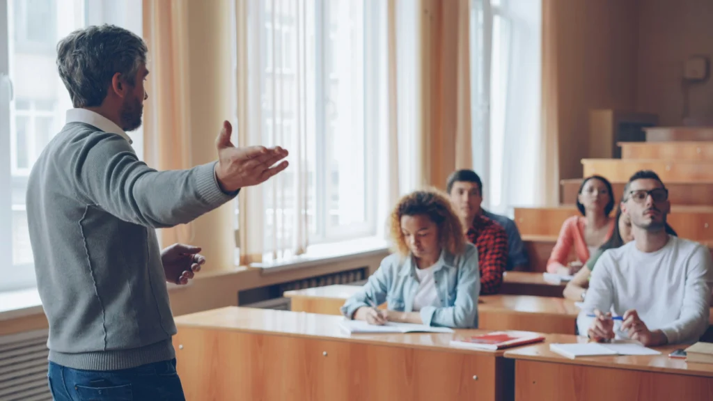 An educator gesturing toward students sitting at desks in a lecture hall, modeling cold-calling strategies using a random name generator wheel.