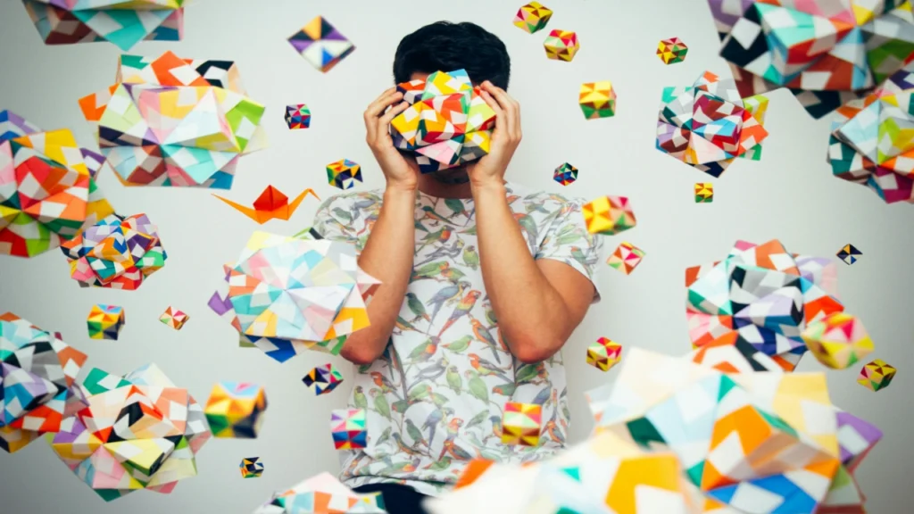 A man holding a complex colorful origami shape over his face while surrounded by floating geometric patterns, showing a chaotic state solved by a random number generator.