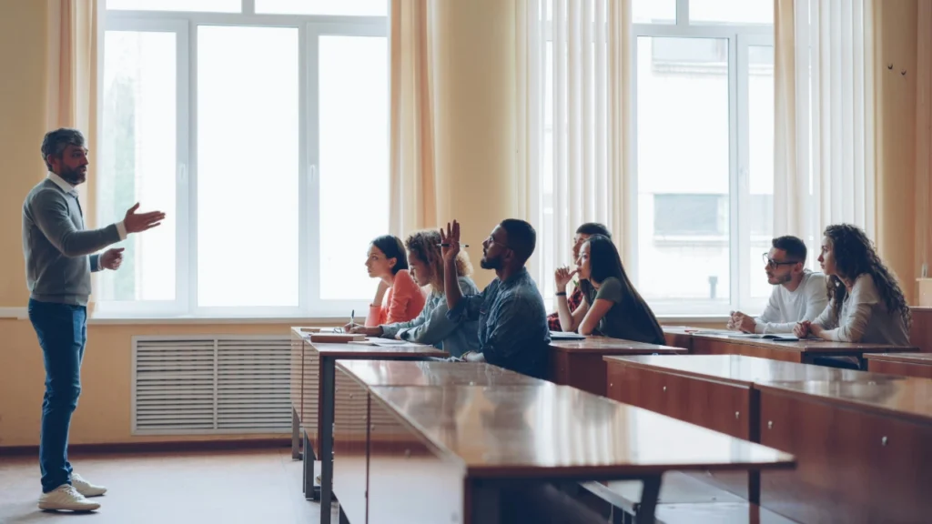 A male teacher lectures a high school class where one student raises a hand, contrasting traditional calling methods with random selection.