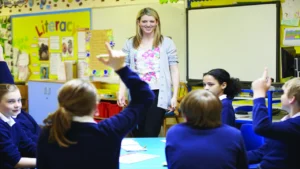A smiling female teacher standing in front of a whiteboard as several school children raise their hands to boost student participation.