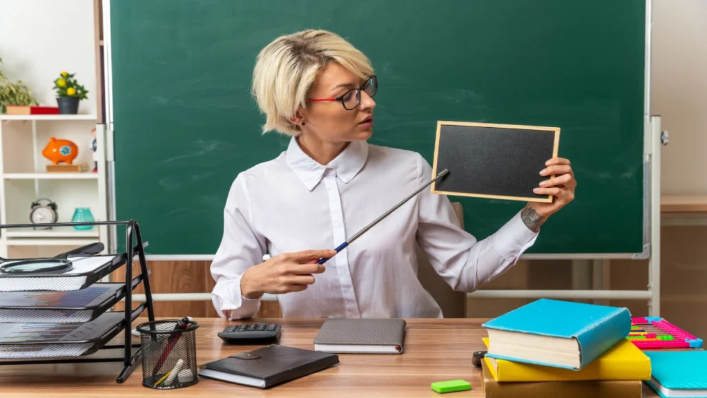 A blonde female teacher with glasses sits at her desk pointing a stick toward a mini blank blackboard to explain a random selection strategy.
