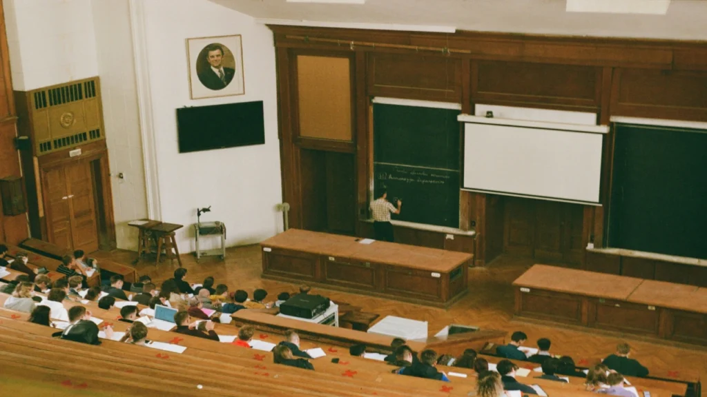 A professor writing on a blackboard using traditional classroom resources in a large university lecture hall filled with students.